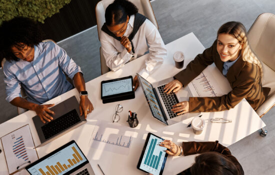 Top view of smiling businesswoman and diverse team discussing graphs and analytics on laptops and tablets at modern office table. Concept of teamwork, digital workflow, and financial strategy.